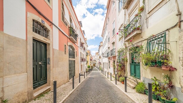 Uma rua de paralelepípedos estreita, ladeada por edifícios com varandas e plantas em vasos.