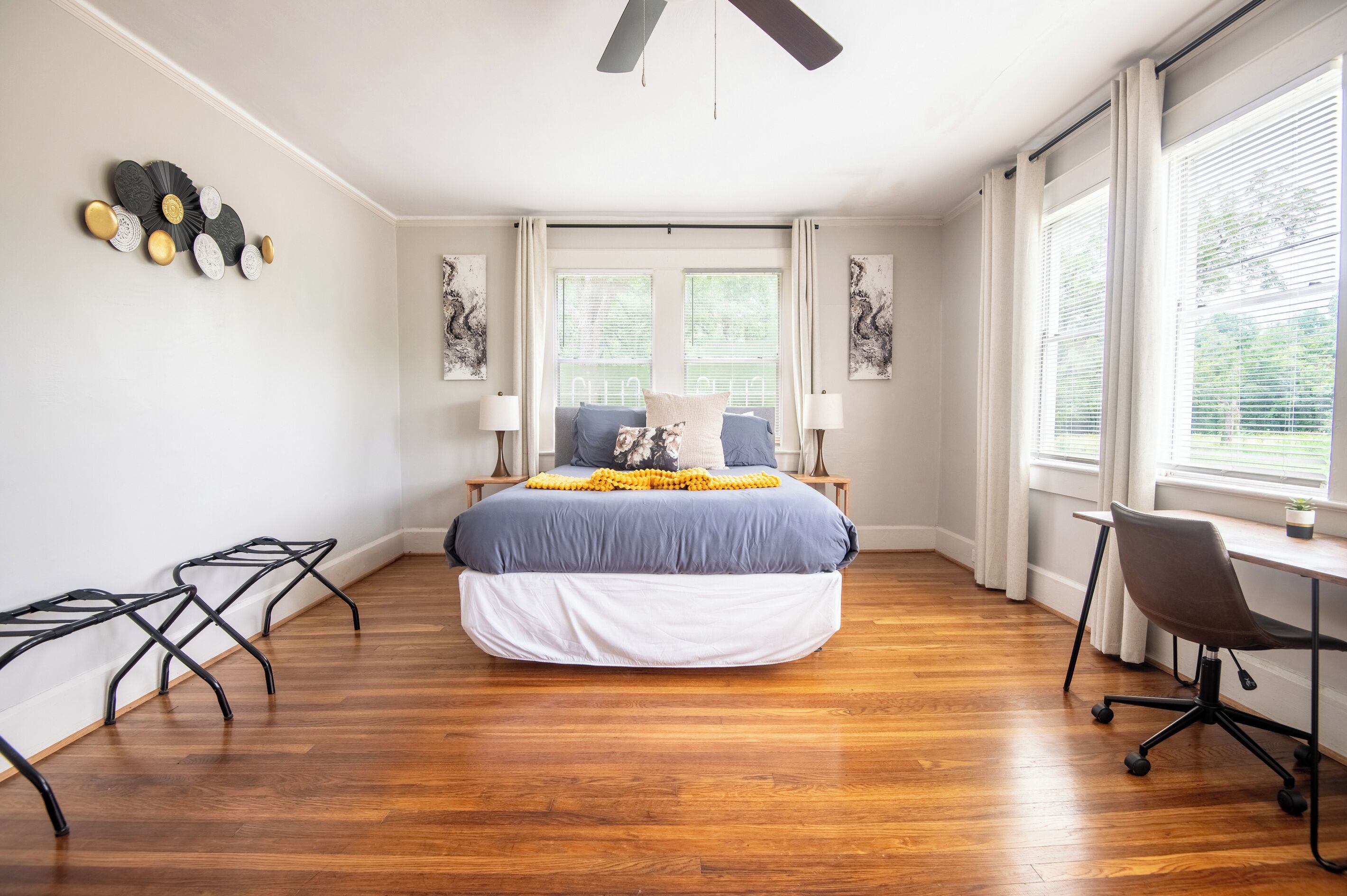 Une chambre à coucher comprenant un lit, deux tables de chevet, un bureau avec une chaise, un ventilateur de plafond et de grandes fenêtres équipées de stores.