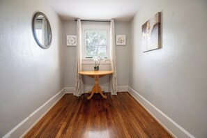 Un pasillo con suelo de madera, una mesa redonda con un jarrón de flores, una ventana con persianas y cuadros en las paredes.