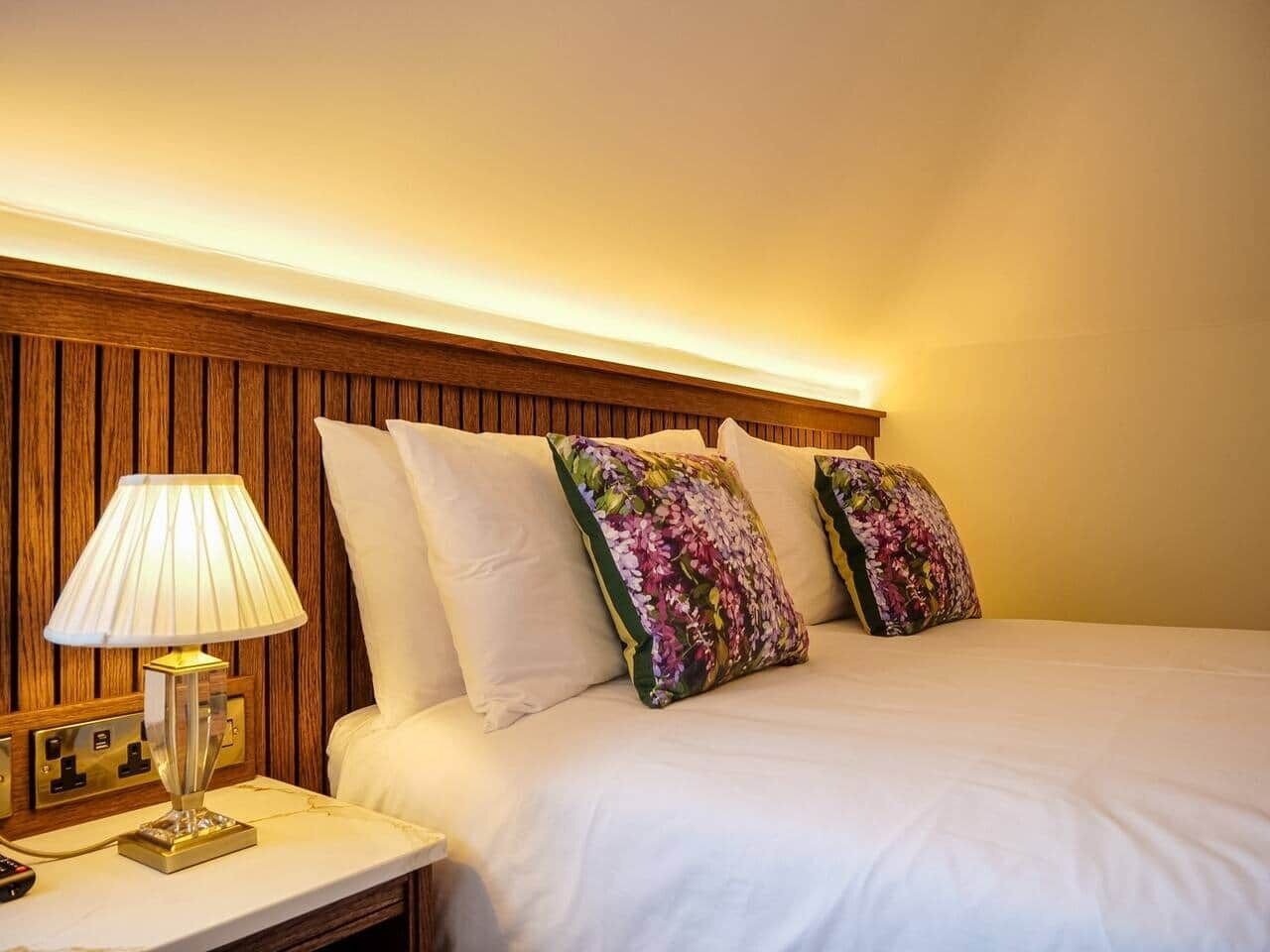 Een hotelkamer met een houten hoofdbord, een bedlampje, een wit bed met bloemenkussens en een nachtkastje.
