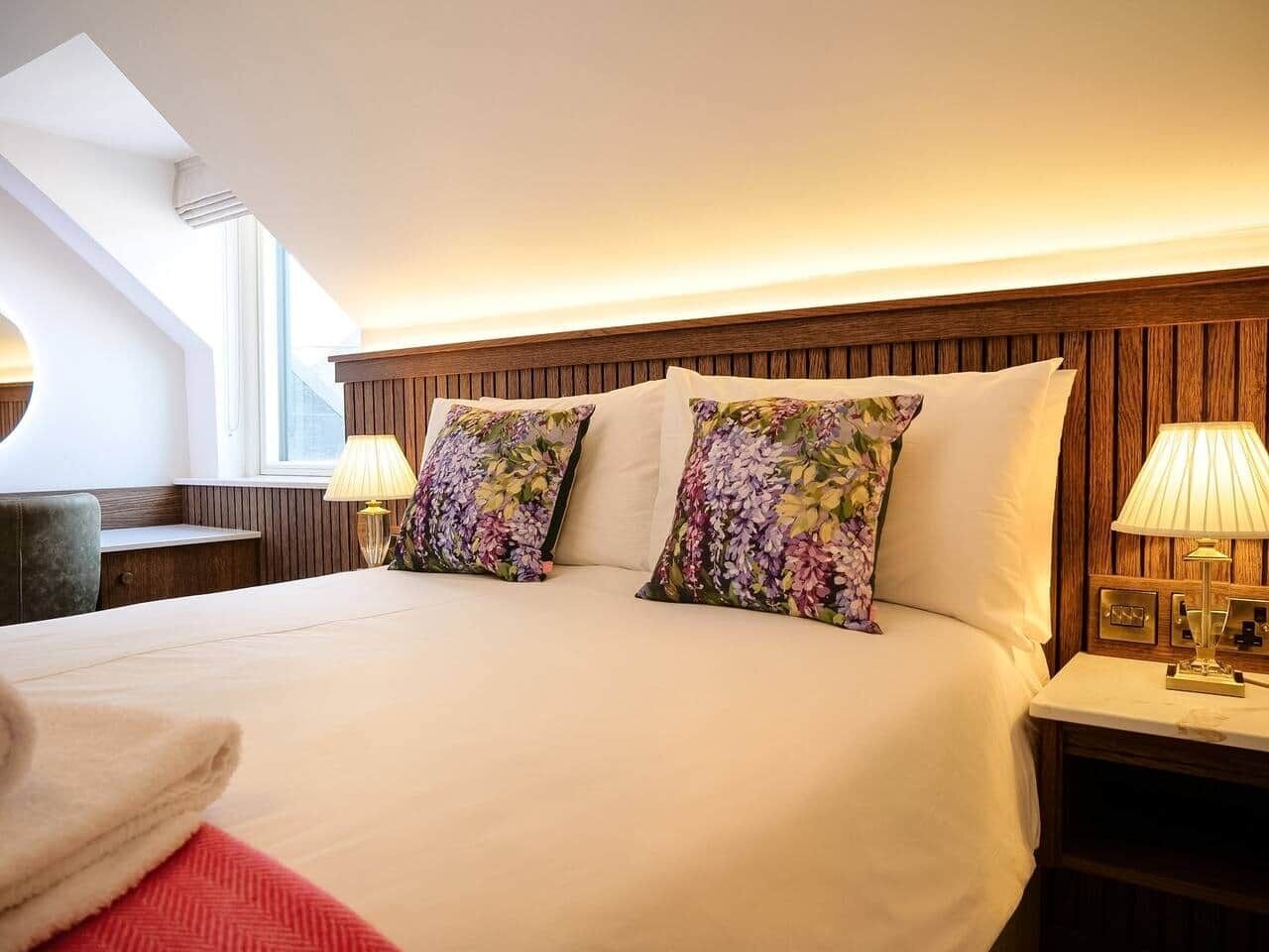 Quarto de hotel com uma cama grande, duas almofadas florais, cabeceira de madeira, luminárias de cabeceira e uma janela com luz natural.