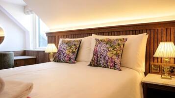 Quarto de hotel com uma cama grande, duas almofadas florais, cabeceira de madeira, luminárias de cabeceira e uma janela com luz natural.