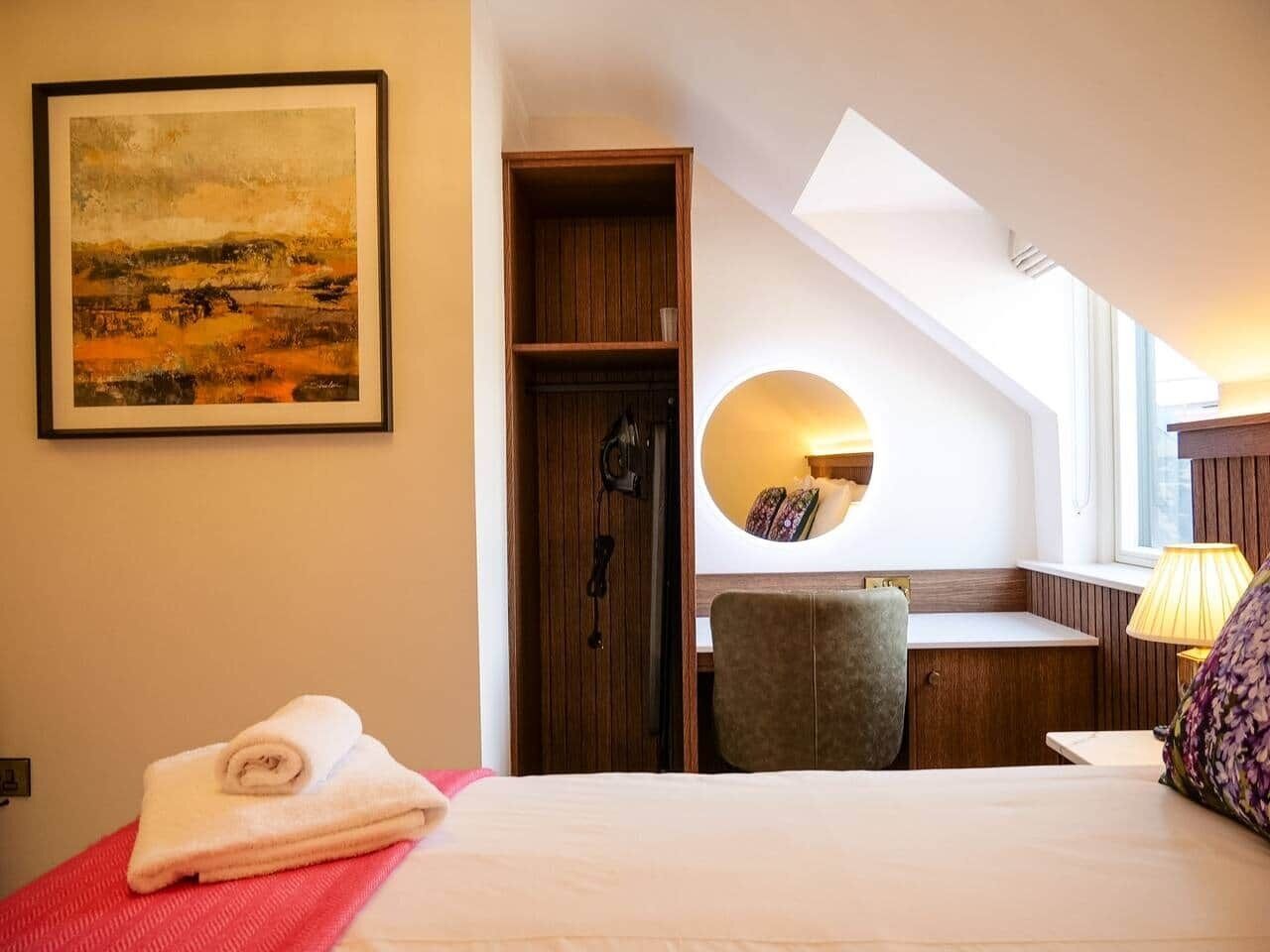 A hotel room with a bed, a desk, a chair, a round mirror, and a painting on the wall.