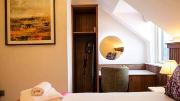A hotel room with a bed, a desk, a chair, a round mirror, and a painting on the wall.