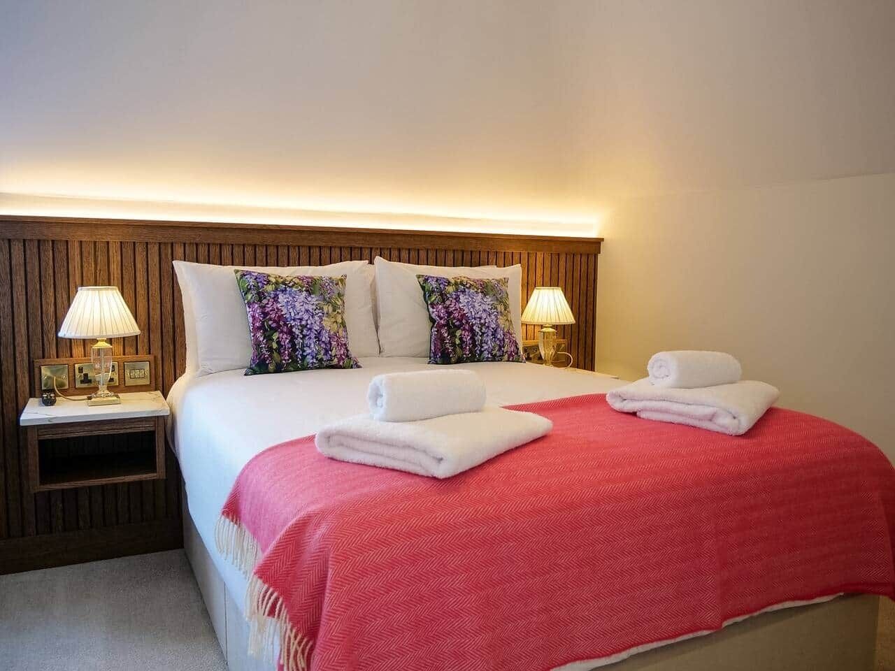 Une chambre d’hôtel avec une tête de lit en bois, deux lampes de chevet, un lit avec une literie blanche et rouge, et deux serviettes enroulées posées sur le lit.