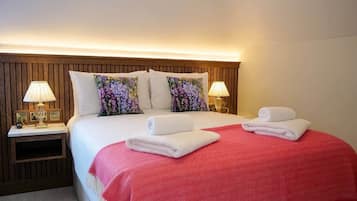 Une chambre d’hôtel avec une tête de lit en bois, deux lampes de chevet, un lit avec une literie blanche et rouge, et deux serviettes enroulées posées sur le lit.