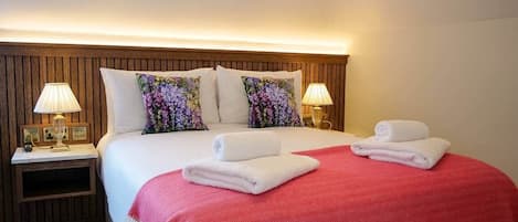 Une chambre d’hôtel avec une tête de lit en bois, deux lampes de chevet, un lit avec une literie blanche et rouge, et deux serviettes enroulées posées sur le lit.