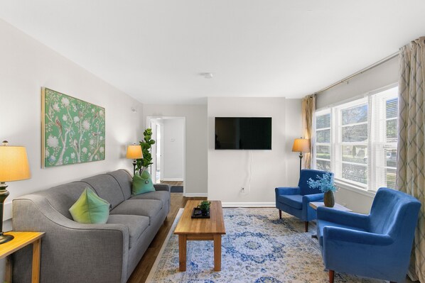 Een woonkamer met een grijze bank, een blauwe fauteuil, een houten salontafel, en een aan de muur bevestigde televisie.