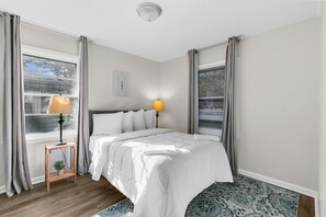 Een slaapkamer met een bed, bedlampen, een raam met gordijnen, een kleintje tafeltje met een plant en een vloerkleed met een patroon.