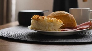 Une part de gâteau sur une assiette, une pâtisserie, une tasse et une bouilloire sur une table près d’une fenêtre donnant sur un paysage verdoyant.
