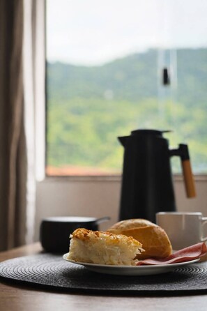 A slice of cake on a plate, a pastry, a cup, and a kettle on a table by a window with a view of greenery. - Casa Vista Verde | Vila do Abraão - Ilha Grande (Angra dos Reis)