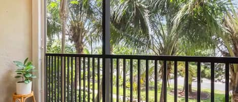 A balcony with a view of tropical vegetation, a wicker chair, a small table with a plant, and a hanging macrame light fixture.