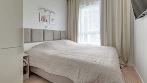 Un dormitorio moderno con cama, mesita de noche, televisor montado en la pared y ventana con cortinas.