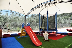Une aire de jeux avec un toboggan, un trampoline et une surface en gazon synthétique.