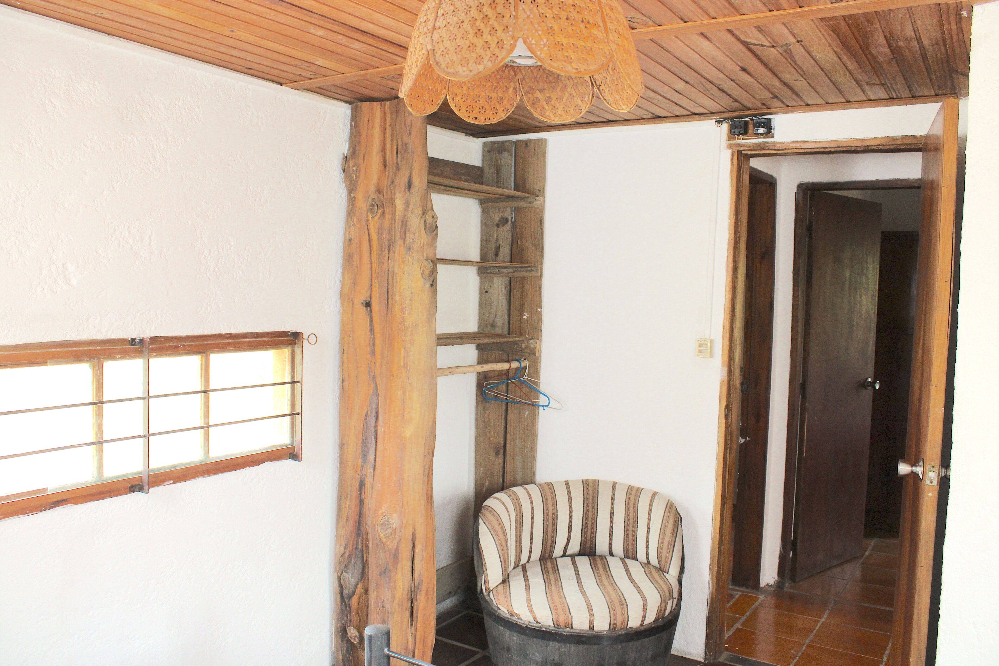 A room with a wooden ceiling, a window with a grid pattern, a striped chair, and a door leading to another room.