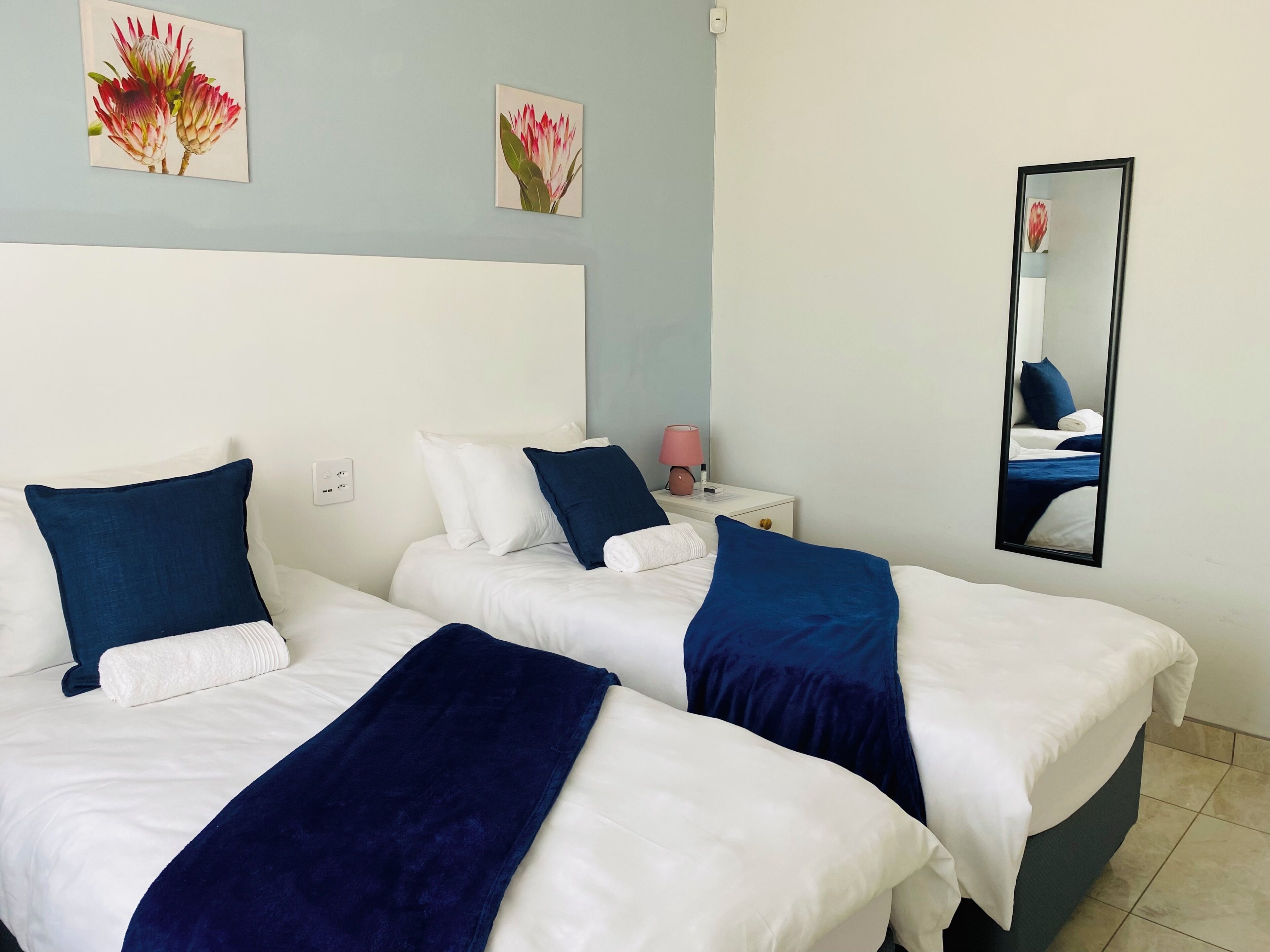 Habitación de hotel con dos camas, ropa de cama blanca, almohadones azules, una mesita de noche con lámpara y obras de arte en las paredes.