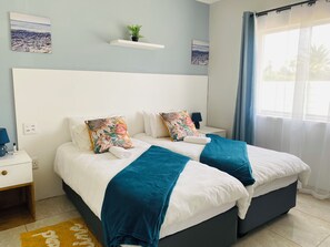 Un lit double avec deux coussins décoratifs, une table de chevet avec une lampe, une petite étagère avec une plante et une fenêtre avec des rideaux.