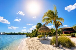 A beach with clear water, a thatched umbrella, palm trees, and a building with a red facade. - Sea View Apartment, Infinity Pool & Beach, 5 min Jan Thiel | B7 (Willemstad)
