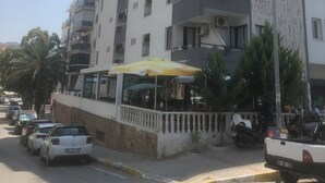 Un immeuble à plusieurs étages doté de balcons, de climatiseurs et situé devant une rue où sont garées des voitures et où se trouve un café.