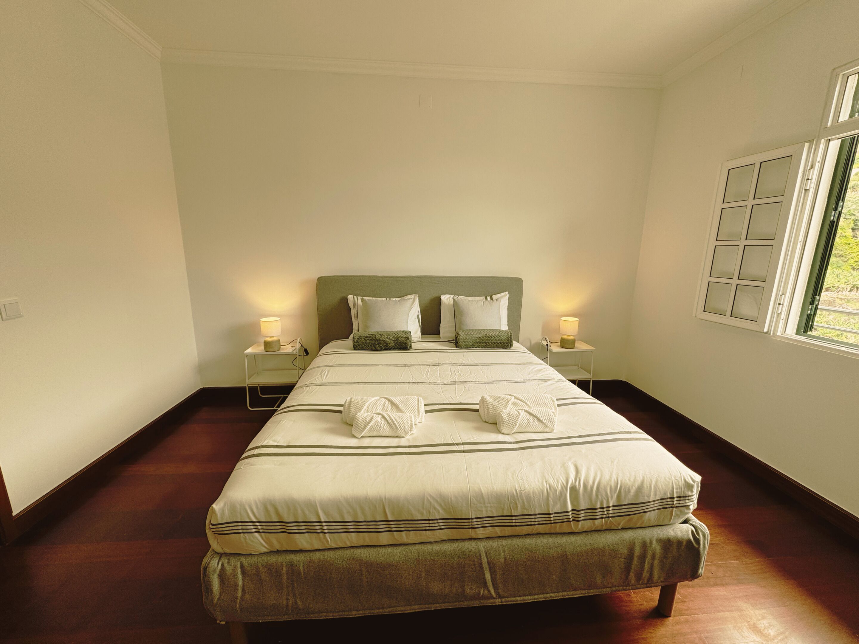 A neatly made bed with pillows, a bedside table with a lamp, and a window with blinds.
