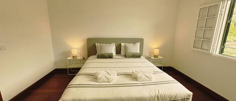 A neatly made bed with pillows, a bedside table with a lamp, and a window with blinds.