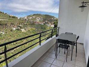 A balcony with a table and chairs, overlooking a hillside with buildings. - Wolf Villa (Câmara de Lobos)