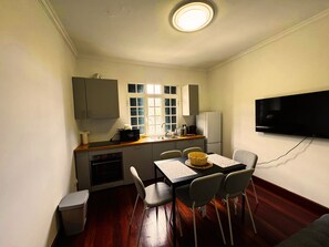 A compact kitchen with a dining table, chairs, a TV, and a window with shutters.