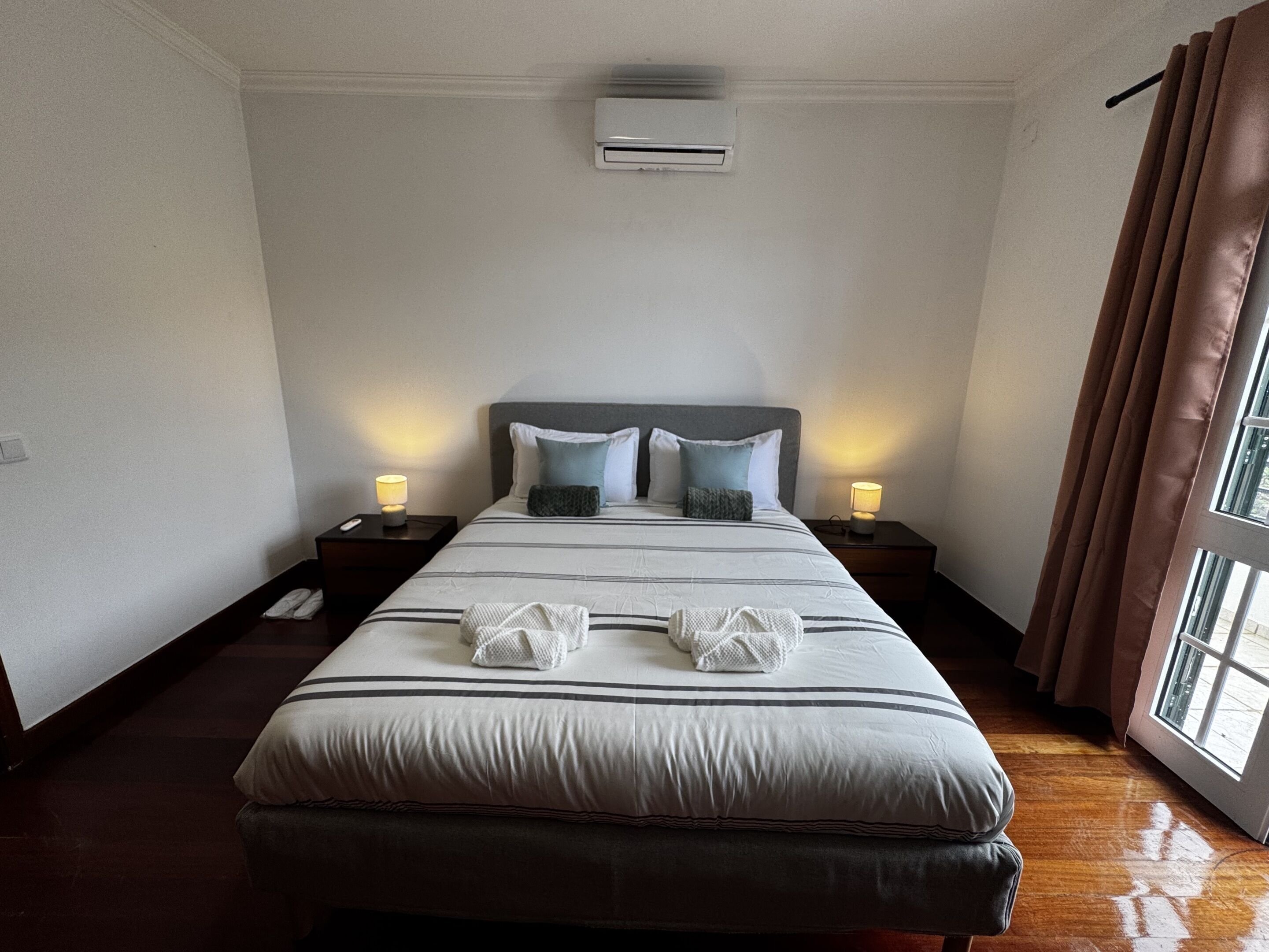 A hotel room with a large bed, two bedside tables with lamps, an air conditioning unit, and a window with curtains.