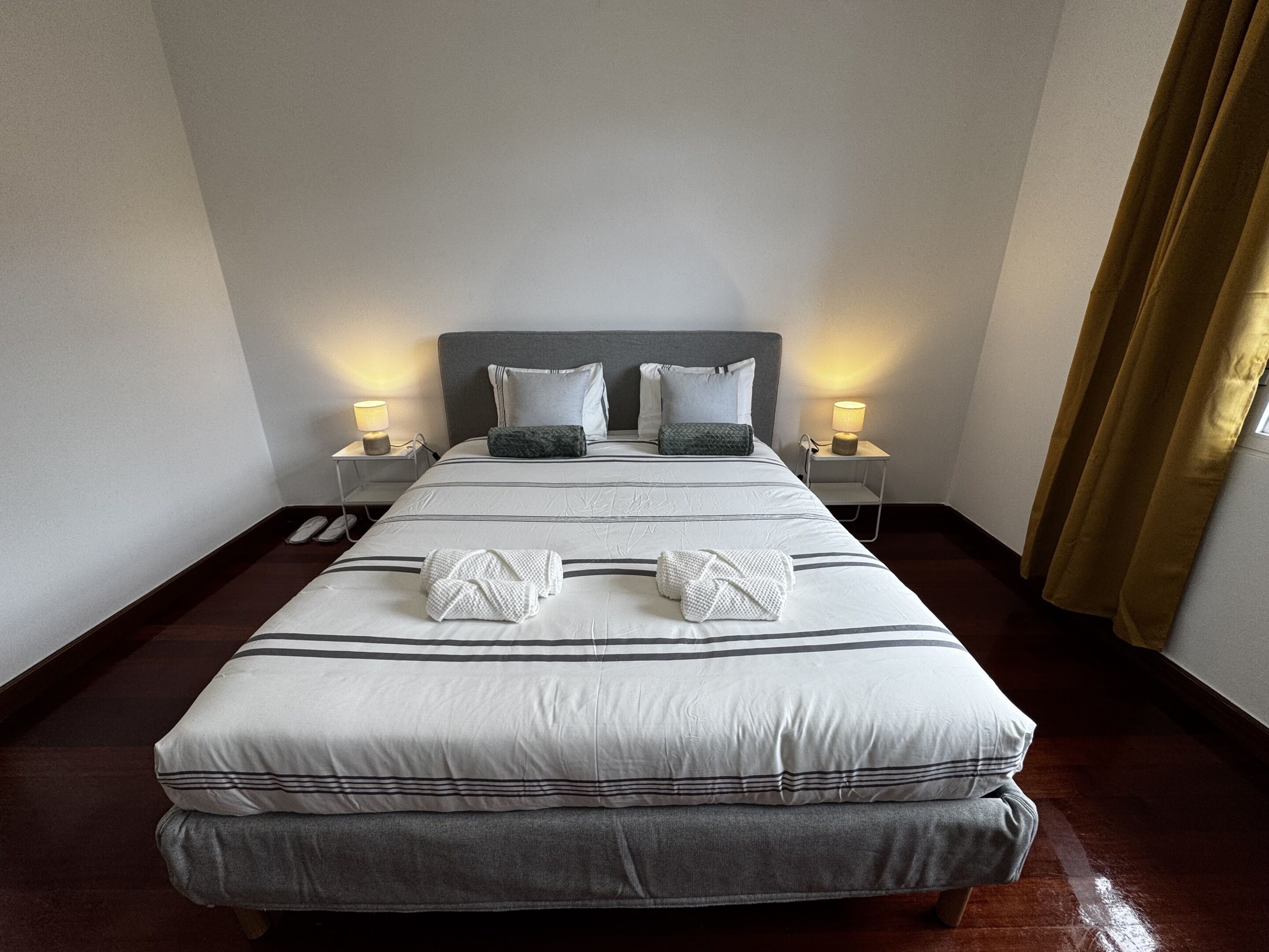Una cama bien hecha con almohadas, una mesita de noche con lámpara y una ventana con cortinas.