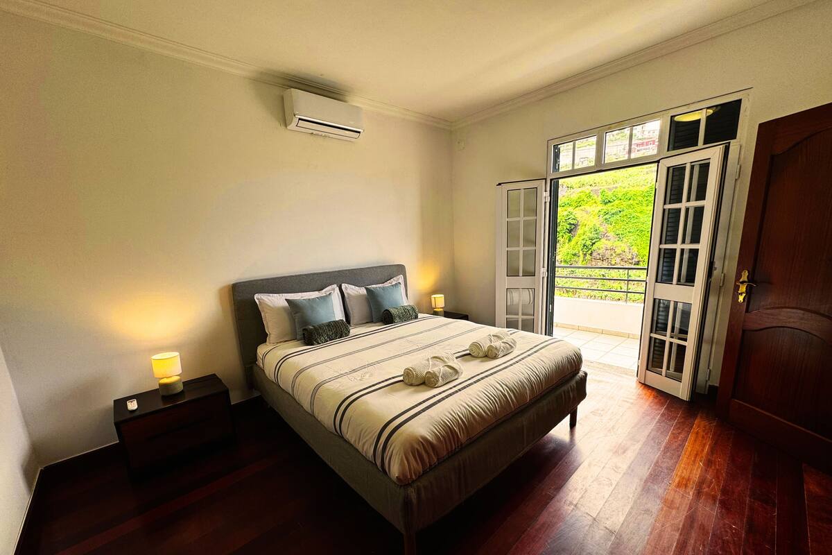 Une chambre à coucher comprenant un lit, des lampes de chevet, un appareil de climatisation, un plancher en bois et une porte donnant sur un balcon.