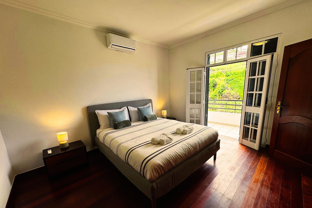 A bedroom with a bed, bedside lamps, an air conditioning unit, a wooden floor, and a door leading to a balcony.