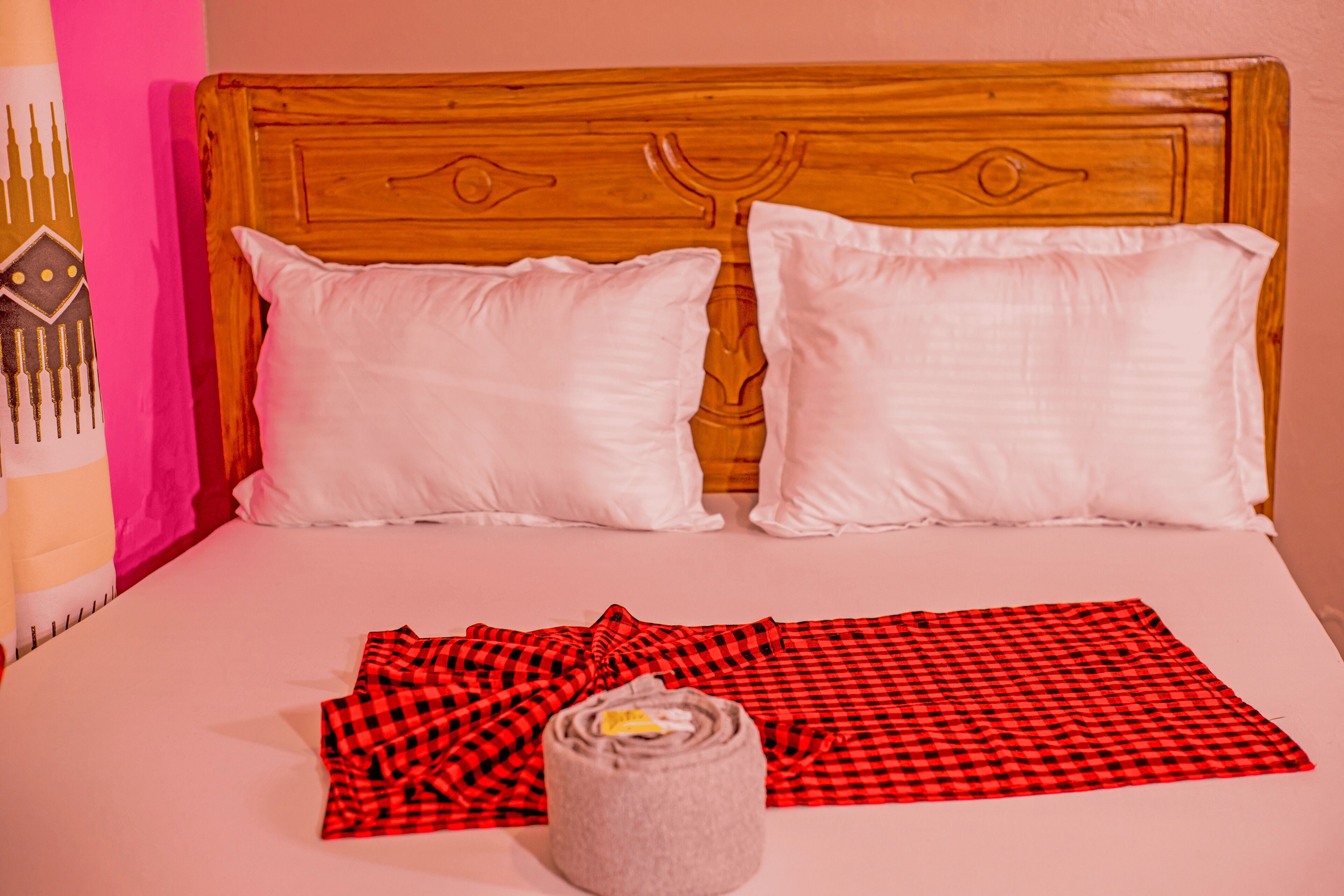 A bed with white pillows, a red and black checkered blanket, and a rolled-up towel.