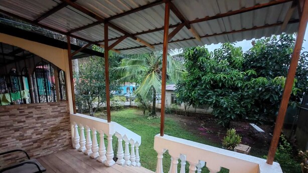 A covered outdoor area with a wooden deck, white balustrades, and a view of a garden with various plants and a blue building in the background. - Congo House. Cottage in Golfito (Golfito)