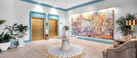A lobby with a round table, two doors, a large abstract painting, and potted plants.