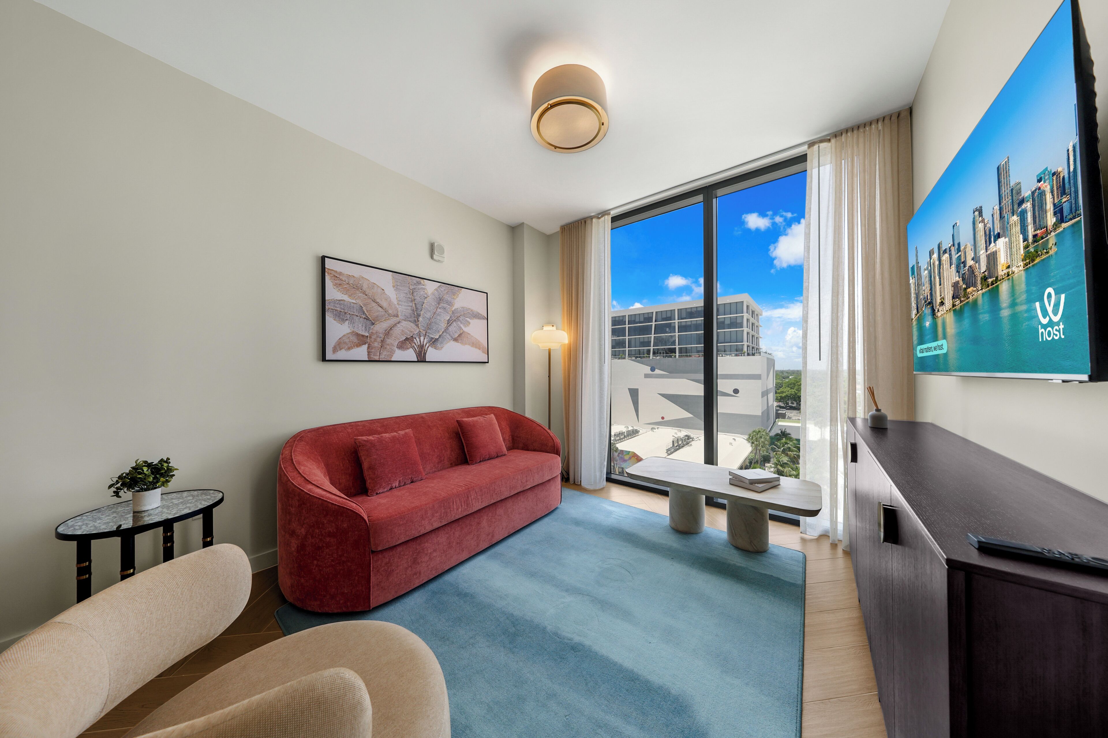 A modern hotel room with a red sofa, a small round table with a plant, a large window with a city view, and a mounted TV on the wall.