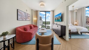A modern hotel room with a red sofa, a beige armchair, a small table, a TV, a bed, and a city view. - Nomad Stlysh (Miami)