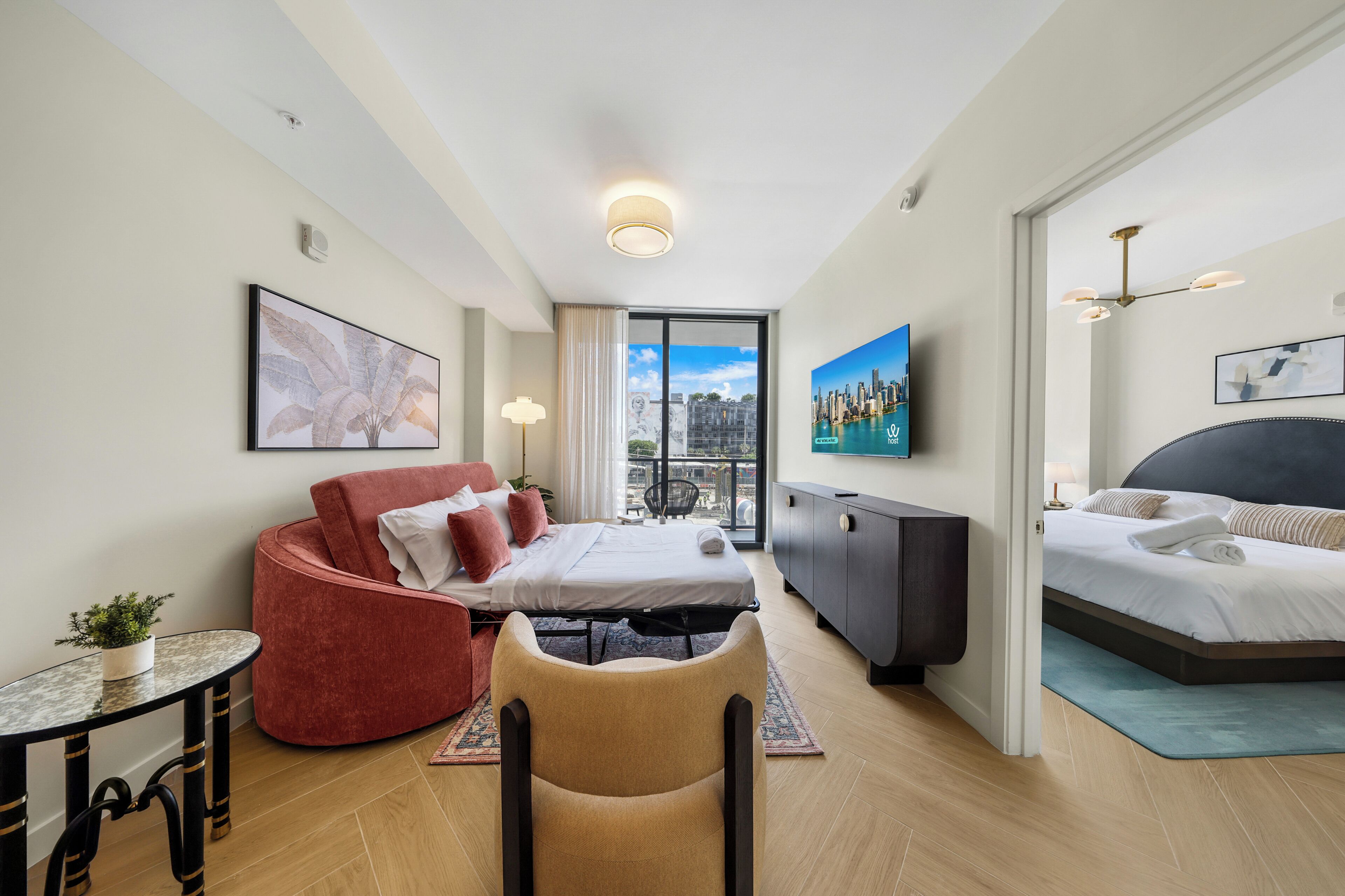 Een moderne hotelkamer met een bed, een rode bank, een klein tafeltje met een plant, een televisie en een stadsgezicht schilderij.