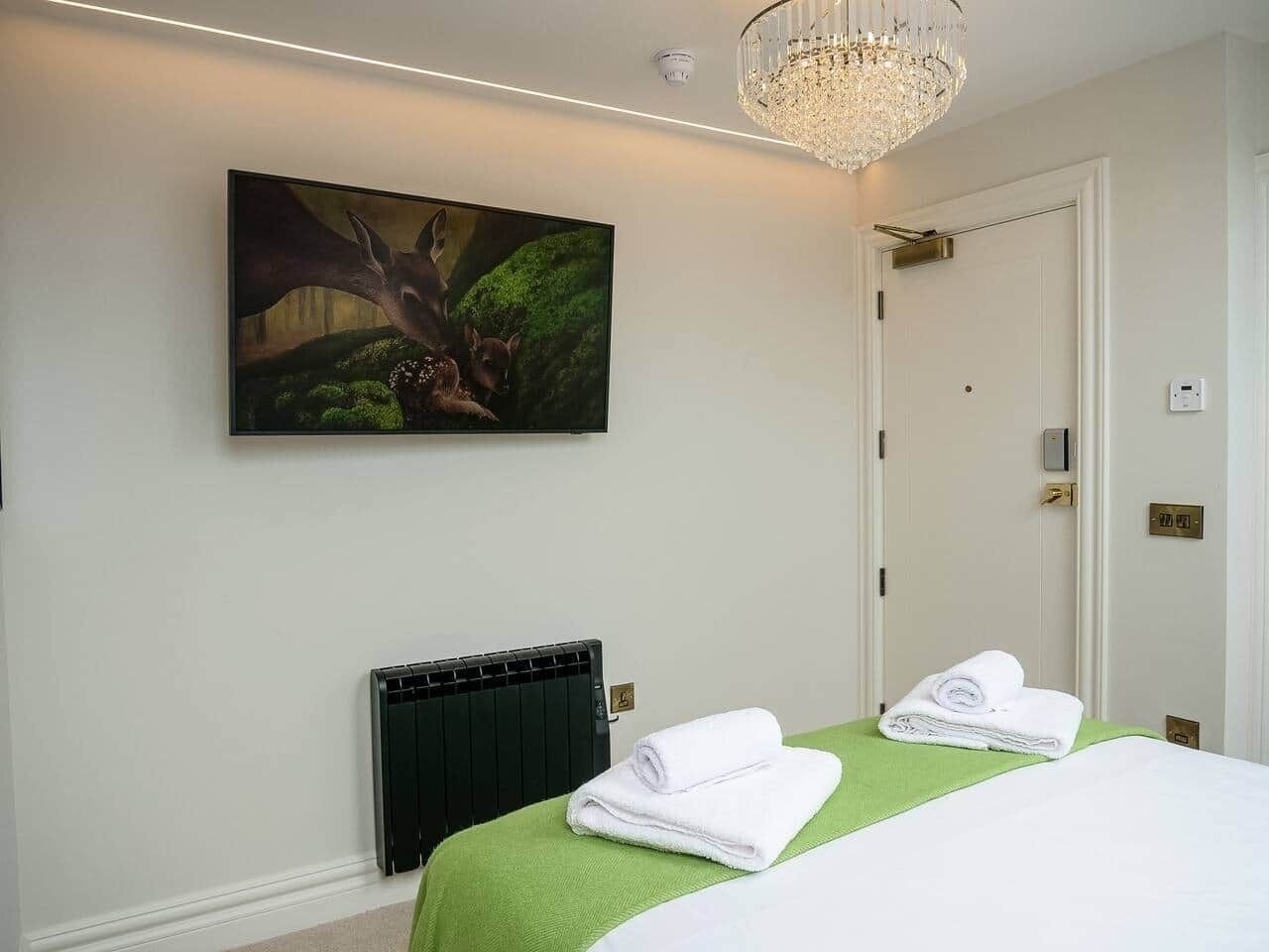 Habitación de hotel con cama, radiador, colcha verde, toalla blanca, un cuadro de ciervos y una araña.