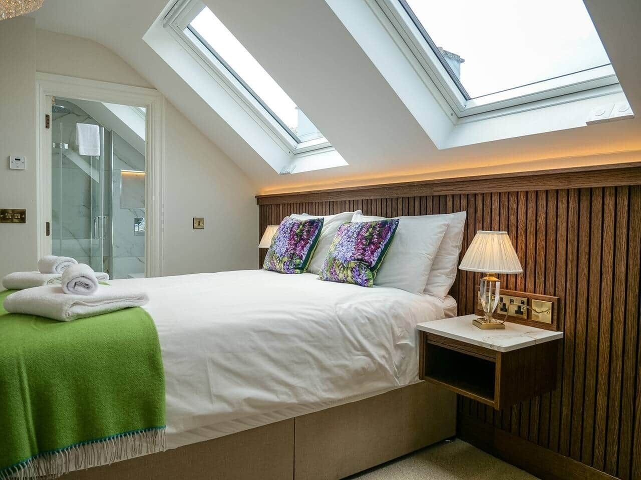 A bedroom with a skylight, a bed with white bedding, a green throw, and a nightstand with a lamp.