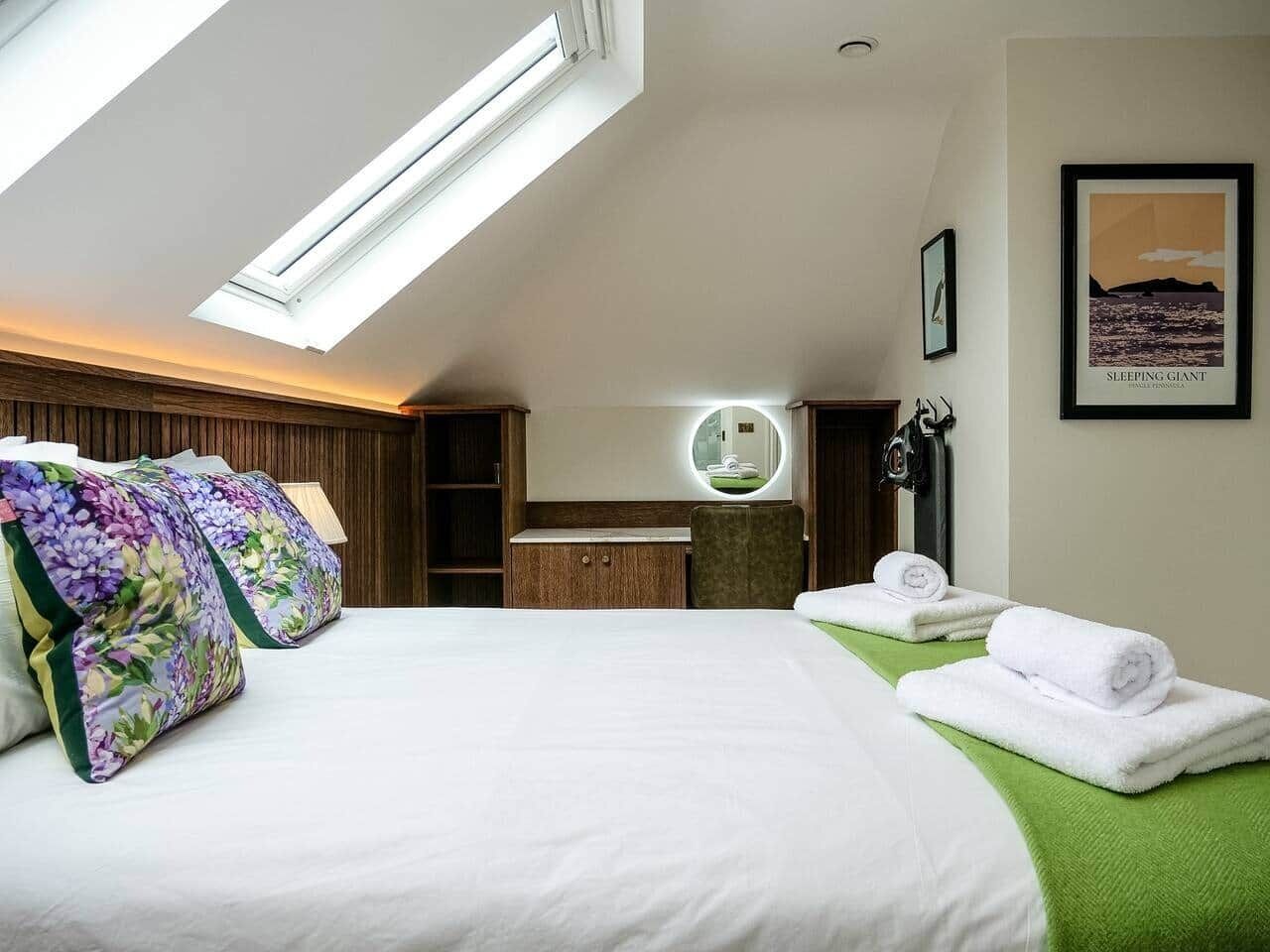 Une chambre d’hôtel avec un puits de lumière, un lit agrémenté d’oreillers à motifs floraux, une coiffeuse avec un miroir et un tableau encadré accroché au mur.