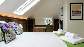 Quarto de hotel com claraboia, cama com almofadas florais, toucador com espelho e quadro pendurado na parede.