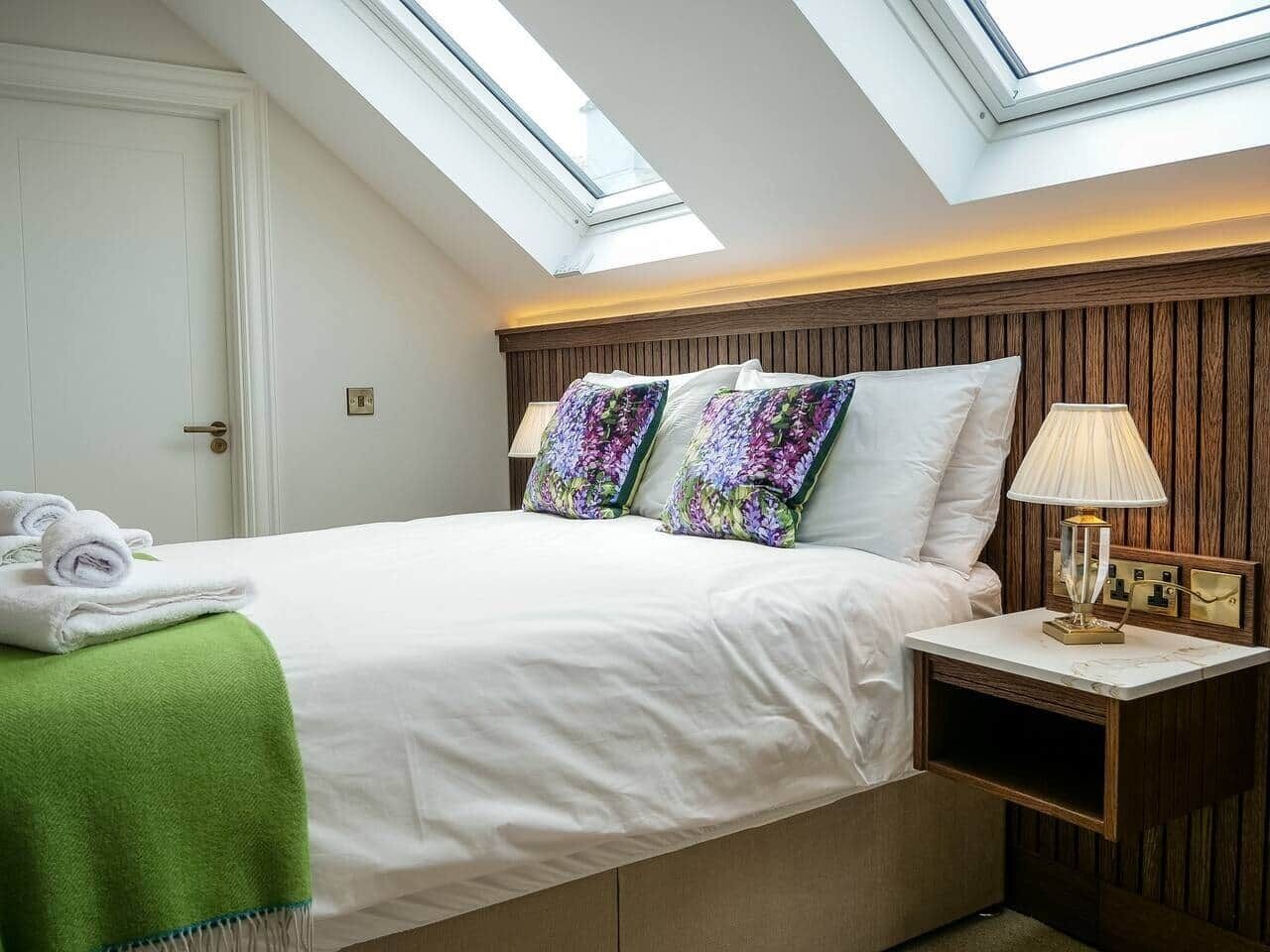 A bedroom with a skylight, a bed with white bedding, two colorful pillows, a green towel, and a bedside lamp.