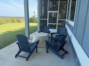 A covered patio with four Adirondack chairs and a round table, overlooking a grassy area and a bridge.
