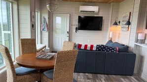 A cozy room with a round wooden table, wicker chairs, a dark sofa with a flag-patterned throw, and a mounted flat-screen TV.