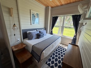 A bedroom with a bed, a nightstand, a wall-mounted light fixture, a window with curtains, and a view of the outdoors.