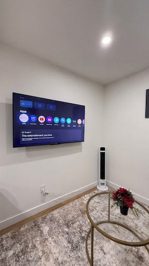 A flat-screen TV mounted on a white wall, a white standing heater, a round metal table with a potted plant, and a marble-patterned carpet. - Our Cozy Corner (Edmonton)