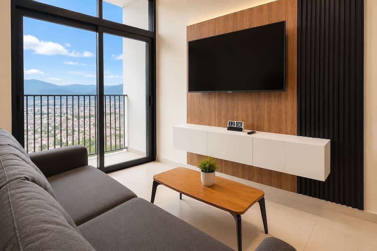 A modern living room with a large window, a flat-screen TV mounted on the wall, a sofa, and a coffee table.