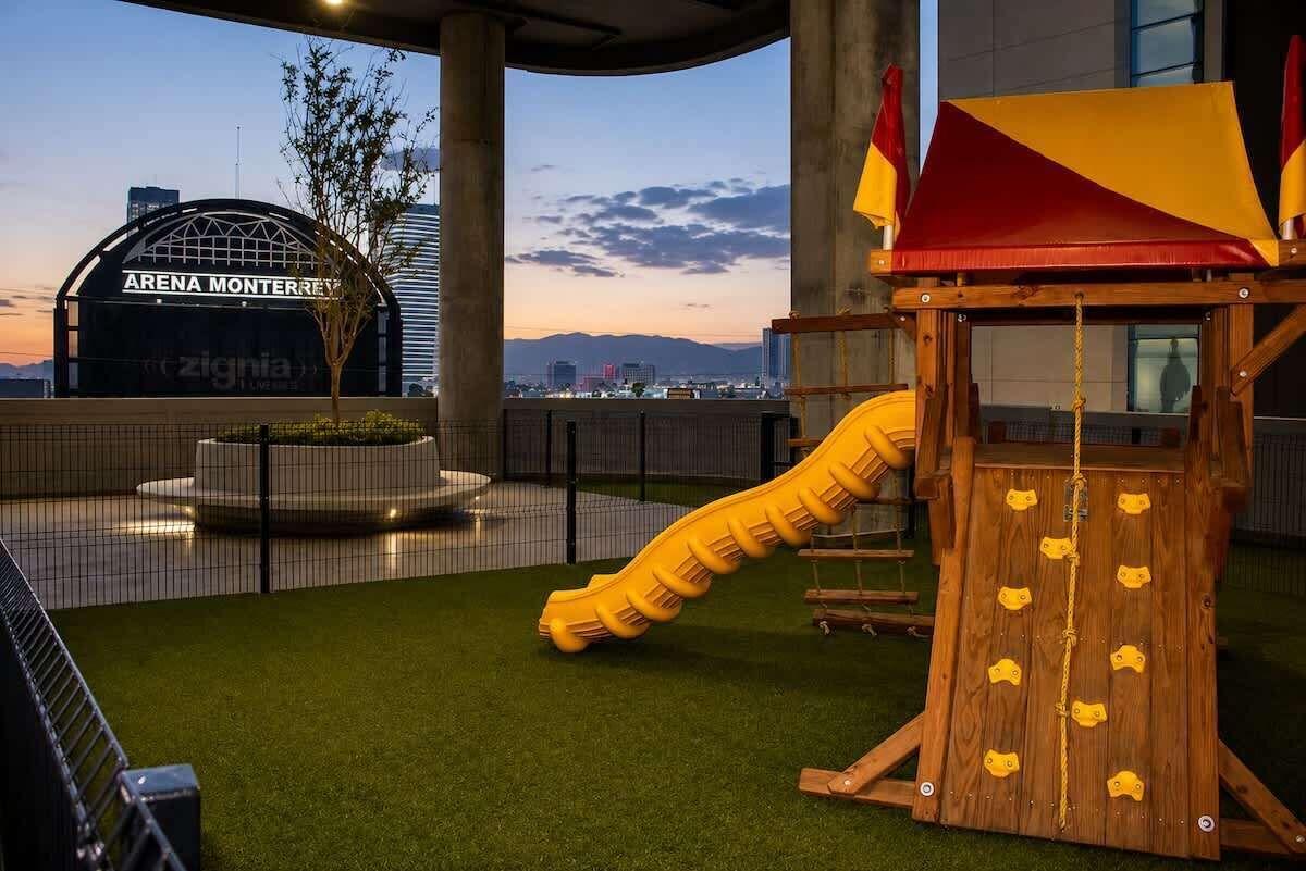 A playground with a slide and climbing structure, surrounded by a fence, with a view of a building labeled 'ARENA MONTERREY' in the background.
