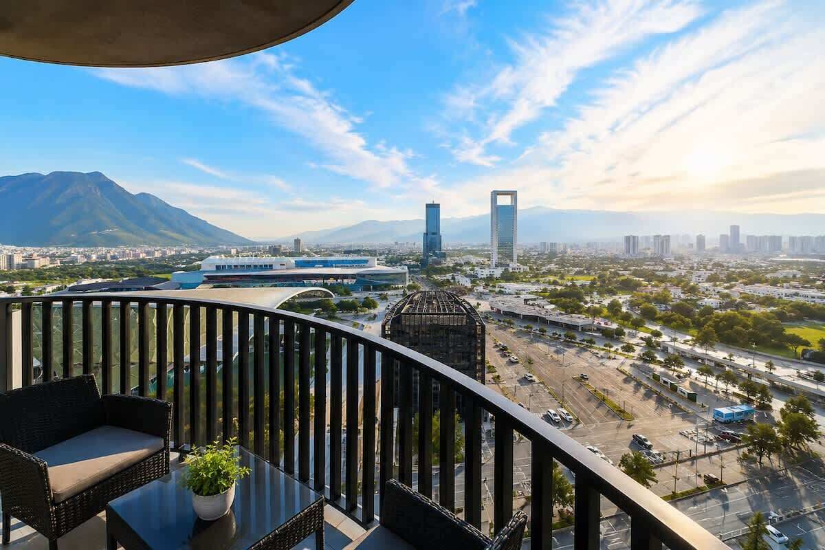 A balcony with wicker furniture, a potted plant, and a view of a cityscape with buildings and mountains.