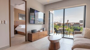A modern hotel room with a bed, a TV, a wooden cabinet, a round coffee table, and a balcony with a view of the city. - Modern Versalles 2BR w/ Rooftop Pool & Views in Puerto Vallarta (Puerto Vallarta)